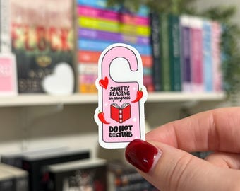 May include: Pink door hanger with a red book and the text "Smutty Reading in progress. Do Not Disturb". The hanger has red chili peppers and hearts on it.