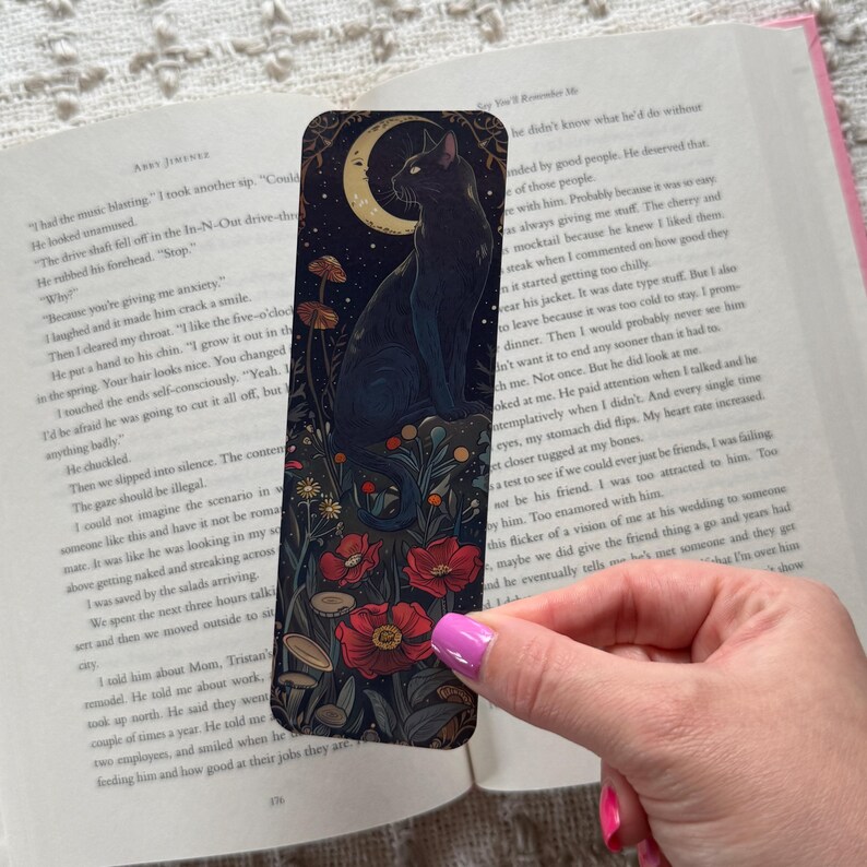 K&ouml;nnte beinhalten: Lesezeichen mit einer schwarzen Katze unter einem Mond, umgeben von Blumen und Pilzen. Das Lesezeichen liegt auf einem offenen Buch.