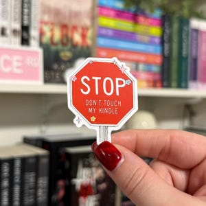 May include: A red stop sign sticker with white text that says "STOP DON'T TOUCH MY KINDLE". The sticker has a white border and is decorated with small pink and white flowers.