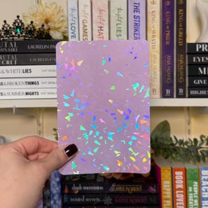 May include: A rectangular, lavender-colored card with a holographic confetti pattern. The card is held in front of a bookshelf filled with books. The confetti reflects various colors, adding a shimmering effect to the card.