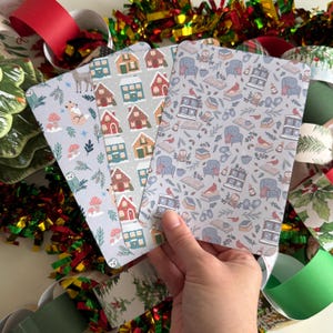 May include: Three rectangular notebooks with different patterns are held in a hand. The notebooks feature illustrations of houses, woodland creatures, and cozy living room scenes. The background includes Christmas decorations with red, green, and gold colors.