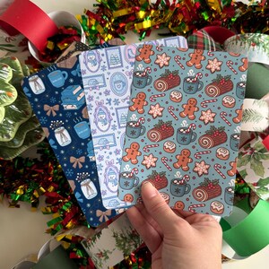 May include: Three decorative notebooks with festive patterns are held in a hand. The notebooks feature designs of snowmen, gingerbread men, mugs of cocoa, and other holiday-themed illustrations. The notebooks are in shades of blue and teal.