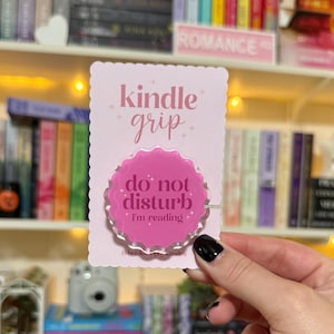 May include: A pink Kindle grip with the text "kindle grip" and "do not disturb I'm reading". The grip has a scalloped edge and is held in front of a bookshelf filled with books.