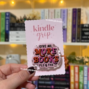 May include: A pink Kindle grip with the words "kindle grip" in cursive and "GIVE ME MORE BOOKS PLS & THX" in a retro font. The grip is held in front of a bookshelf.