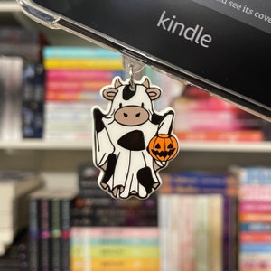 May include: A white and black cow dressed as a ghost holding a small orange pumpkin. The cow is wearing a white sheet with black spots and has a black and white face. The cow is holding the pumpkin in its right hand. The cow is hanging from a black and silver Kindle e-reader.