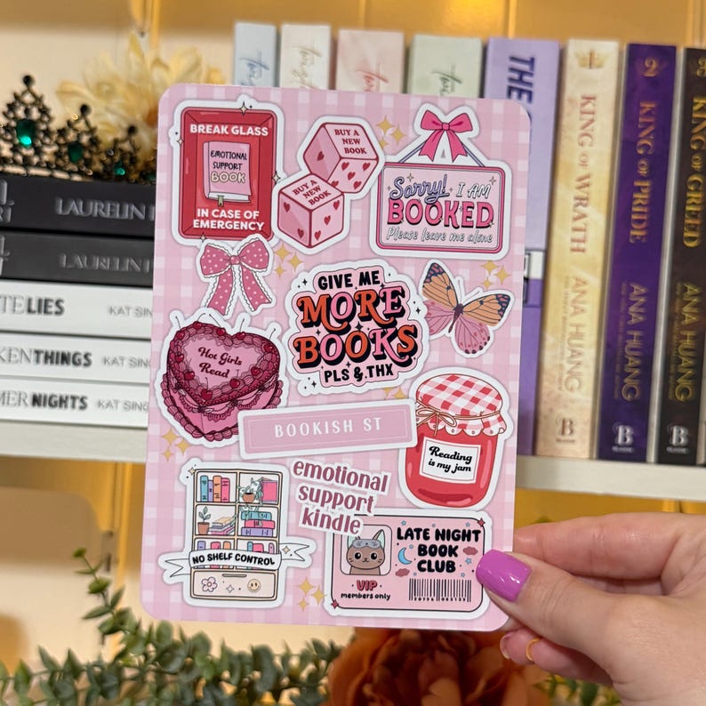 May include: A pink sticker sheet with various book-themed designs. Includes phrases like "Give Me More Books," "Emotional Support Book," and "Late Night Book Club." Features bows, dice, a butterfly, and a jar of jam.