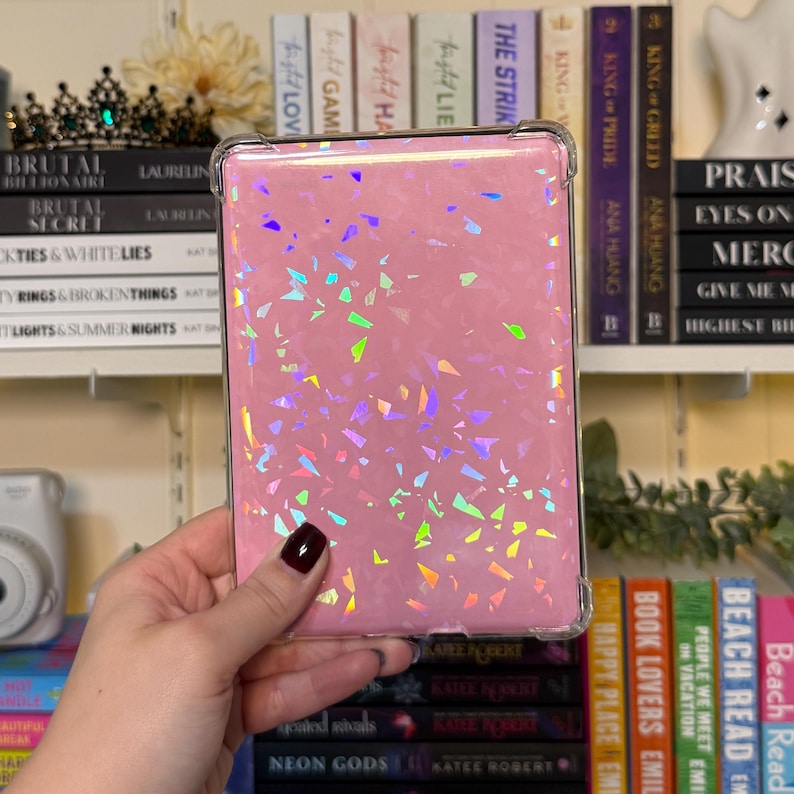 May include: A pink e-reader case with a clear, protective bumper. The case is covered in iridescent, confetti-like shapes. The e-reader is held in a hand, with a shelf of books in the background.