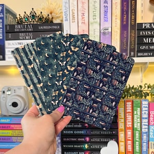 May include: A hand holds a stack of decorative book covers with various patterns. Designs include butterflies, moons, ferns, and a bookshelf motif. The covers are in shades of teal, navy, and gold, with a soft, matte finish. The covers are for books.