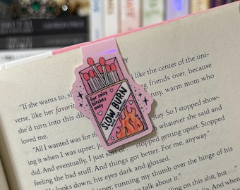 Puede incluir: Un marcapáginas rosa con un diseño de caja de cerillas. La caja de cerillas tiene el texto "SLOW BURN book club matches" y "for spicy bitches only". Las cerillas son rojas y las llamas son naranjas y amarillas. El marcapáginas está en un libro.