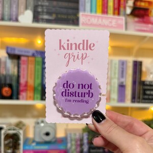 May include: A light purple Kindle grip with the text "kindle grip" in pink script. The grip has a scalloped edge and the text "do not disturb I'm reading" in white. The grip is on a pink scalloped card.