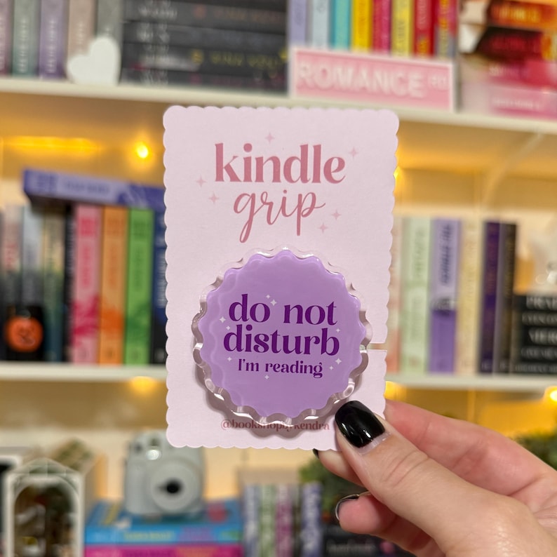 May include: A light purple Kindle grip with the text "kindle grip" in pink script. The grip has a scalloped edge and the text "do not disturb I'm reading" in white. The grip is on a pink scalloped card.