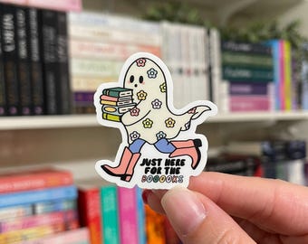May include: A white ghost with colorful floral patterns is carrying a stack of books. The ghost is wearing pink cowboy boots and blue jeans. The text "Just here for the booooks" is written below the ghost.