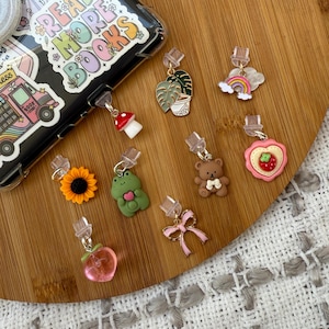 May include: A collection of 10 colorful phone charms, including a sunflower, a frog, a mushroom, a potted plant, a rainbow, a peach, a teddy bear, a cookie, a bow, and a book charm. The charms are attached to clear plastic clips.