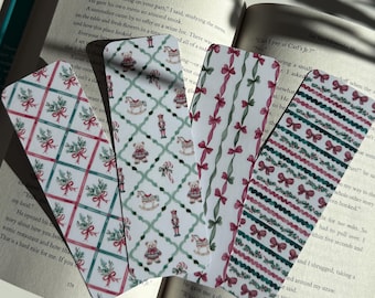 May include: Four decorative bookmarks with festive designs. One features a grid pattern with red bows and greenery, another has a repeating pattern of teddy bears and rocking horses, and the other two have bow and stripe patterns. The bookmarks are resting on an open book.