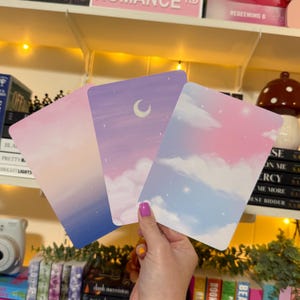 May include: Three rectangular art prints featuring pastel sky scenes. One print has a pink and blue gradient, another depicts a moon and clouds, and the third shows clouds in pink and blue. The prints are held by a person.