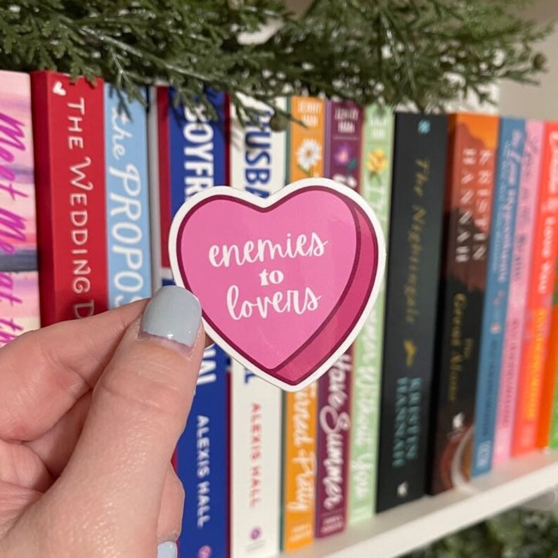 K&ouml;nnte beinhalten: Ein rosa herzf&ouml;rmiger Aufkleber mit den Worten "enemies to lovers" in wei&szlig;er Schrift. Der Aufkleber wird vor einem B&uuml;cherregal mit bunten B&uuml;chern gehalten. Der Aufkleber hat einen dunkelrosa Rand.