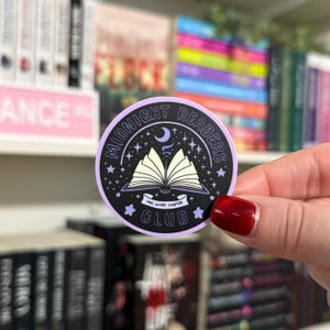 May include: A purple and black round sticker with the text "Midnight Readers Club" and an illustration of an open book with stars and a crescent moon. The text "One More Chapter" is written on a banner below the book.