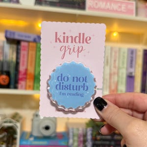 May include: A light pink Kindle grip with the words "kindle grip" in pink script. The grip has a light blue, scalloped-edge circle with the words "do not disturb, I'm reading" in white. The grip is on a pink card.