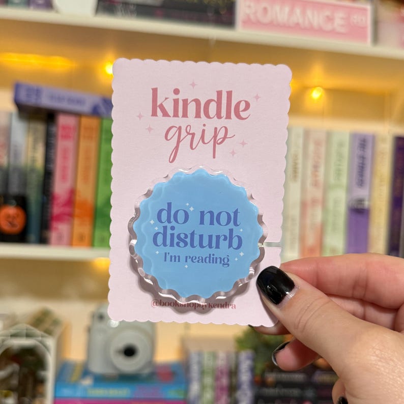 May include: A light pink Kindle grip with the words "kindle grip" in pink script. The grip has a light blue, scalloped-edge circle with the words "do not disturb, I'm reading" in white. The grip is on a pink card.