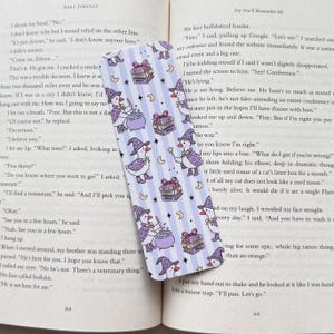 May include: A rectangular bookmark featuring a repeating pattern of white ducks wearing purple witch hats and capes, casting spells with wands over purple cauldrons, interspersed with stacks of books tied with bows and small yellow crescent moons.
