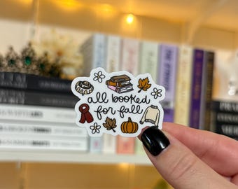 May include: A white sticker with the text "all booked for fall" and fall-themed illustrations. The sticker features a cinnamon roll, books, a leaf, a sweater, a pumpkin, and flowers. The sticker is held by a person.