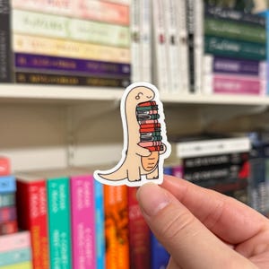 May include: A beige dinosaur sticker holding a stack of colorful books. The sticker is on a hand against a background of bookshelves.