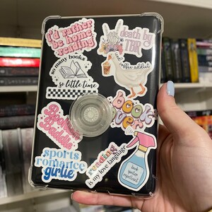 K&ouml;nnte beinhalten: Ein schwarzer E-Reader mit einer transparenten Schutzh&uuml;lle, bedeckt mit bunten Aufklebern. Aufkleber zeigen S&auml;tze wie "I'd rather be home reading" und "Reading is my love language". Ein klarer, kreisf&ouml;rmiger Handyhalter ist angebracht.