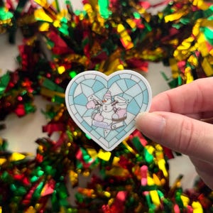 May include: A heart-shaped sticker featuring a winter scene with a snowman, mittens, and boots. The design is in a stained-glass style with a light blue and white color scheme. The word "Winter" is written on the bottom of the sticker.