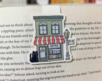 Puede incluir: Una pegatina de un edificio verde con un toldo rojo y blanco. El edificio tiene un letrero que dice "Book Tales" y un gato negro está sentado frente a él. La pegatina está en una página de un libro.