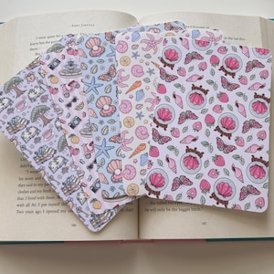 May include: A set of five rectangular bookmarks featuring various repeating patterns including pink seashells, butterflies, strawberries, books, trees, cameras, and scooters, all displayed in a fan arrangement against an open book background.
