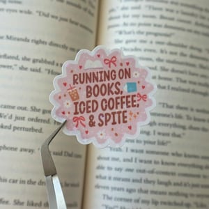 May include: A scalloped, light pink sticker featuring the brown text RUNNING ON BOOKS, ICED COFFEE, & SPITE. The design includes small illustrations of a book, a bow, a cup of iced coffee, a blue square, and tiny red hearts and white flowers.