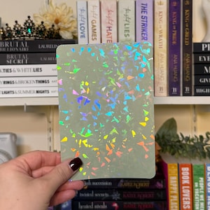 May include: A rectangular, pale green card with a holographic, multicolored confetti pattern. The card is held in front of a bookshelf filled with books. The books have titles such as "Brutal Billionaire" and "King of Greed".