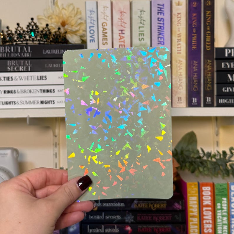 May include: A rectangular, pale green card with a holographic, multicolored confetti pattern. The card is held in front of a bookshelf filled with books. The books have titles such as "Brutal Billionaire" and "King of Greed".