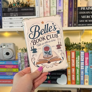 May include: Clear phone case featuring the words "Belle's Book Club" with a rose in a glass dome design. The case has a vintage aesthetic with a star and decorative elements. The case is held in a hand in front of a bookshelf.