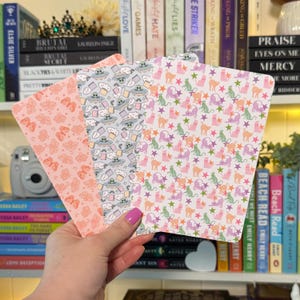 May include: Three patterned paper sheets are held in a hand. The sheets feature repeating designs of butterflies, UFOs with aliens, and cats with stars. The color palette includes pink, gray, green, and orange.