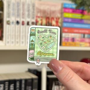 May include: A green and white sticker of a book with a heart-shaped map of a small town. The book cover says "Small Town Romance".
