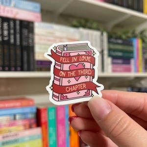 May include: A pink and red sticker with a book illustration and the text "Fell in love on the third chapter".
