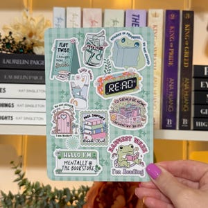 May include: A sticker sheet with various book-themed illustrations. Designs include a frog reading, a stack of books, a clock that says "RE:AD", and phrases like "I'd rather be home" and "mentally @ the bookstore". The background is a light green checkered pattern.