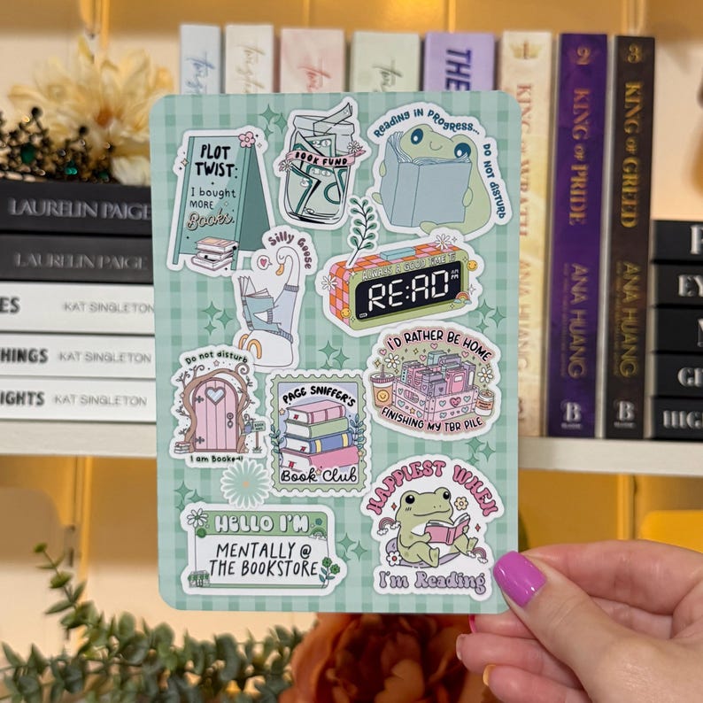 May include: A sticker sheet with various book-themed illustrations. Designs include a frog reading, a stack of books, a clock that says "RE:AD", and phrases like "I'd rather be home" and "mentally @ the bookstore". The background is a light green checkered pattern.