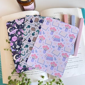 May include: Three decorative book covers with unique patterns. One features a dark purple floral design with a moth, another has swords and potions, and the last has roses and books. Highlighters and a candle are also visible.