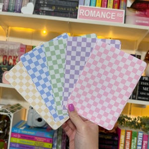 May include: A hand holds five rectangular notepads with a checkered pattern in pastel colors: yellow, blue, green, purple, and pink. The notepads are displayed in front of a bookshelf with the text "ROMANCE RD" visible.
