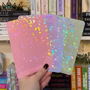 May include: A hand holding four rectangular sheets with a holographic, confetti-like design. The sheets are in shades of pink, lavender, and green. Bookshelves and book titles are visible in the background.