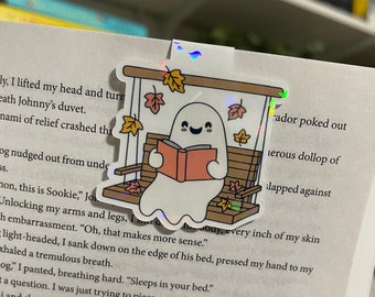 May include: A holographic sticker featuring a white ghost sitting on a wooden swing with a red book. The ghost is smiling and has a happy expression. The swing is decorated with autumn leaves. The sticker is perfect for adding a touch of whimsy to your planner, journal, or laptop.