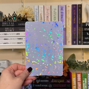 May include: A rectangular, light-colored card with a holographic, confetti-like pattern of various colors. The card is held in front of a bookshelf filled with books. The holographic design reflects shades of blue, green, pink, and yellow.