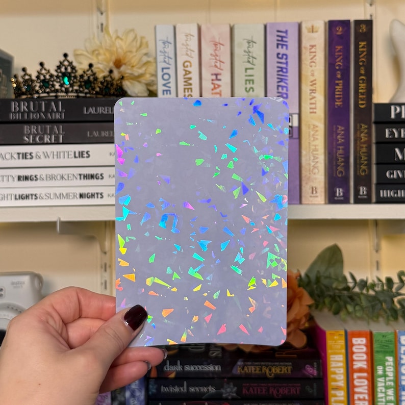 May include: A rectangular, light-colored card with a holographic, confetti-like pattern of various colors. The card is held in front of a bookshelf filled with books. The holographic design reflects shades of blue, green, pink, and yellow.