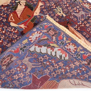 May include: A detailed shot of a Persian rug with a deep blue field and intricate patterns. The rug features figures playing instruments and floral motifs in red, brown, and beige. The rug's border is a lighter blue with repeating designs.