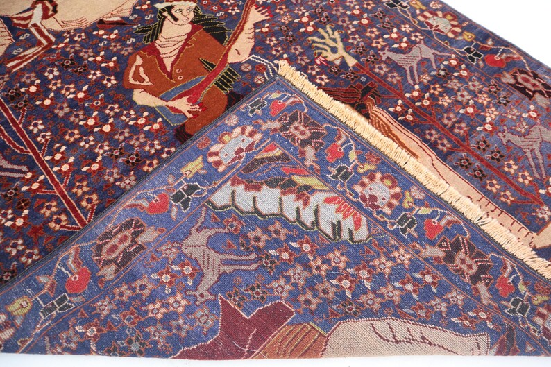 May include: A detailed shot of a Persian rug with a deep blue field and intricate patterns. The rug features figures playing instruments and floral motifs in red, brown, and beige. The rug's border is a lighter blue with repeating designs.