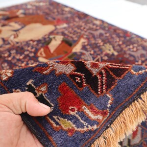 May include: A detailed, hand-knotted rug with a rich, dark blue background and a complex pattern. The design features figures in shades of brown, red, and beige, with a border of intricate geometric shapes. The rug has a fringe of tan-colored threads.