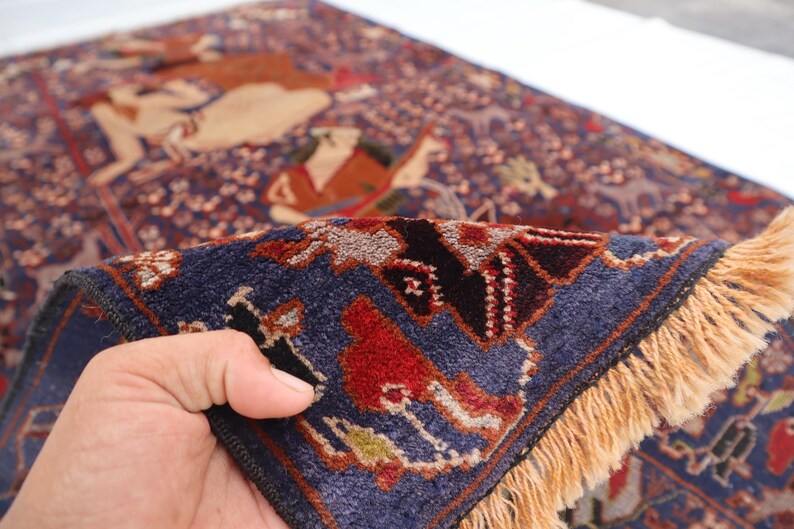 May include: A detailed, hand-knotted rug with a rich, dark blue background and a complex pattern. The design features figures in shades of brown, red, and beige, with a border of intricate geometric shapes. The rug has a fringe of tan-colored threads.