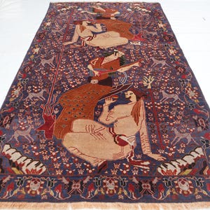 May include: A rectangular, hand-woven rug with a deep blue background and intricate patterns. The rug features figures in beige tones, deer, and floral motifs. The border is a mix of red, beige, and blue, with a fringed edge.